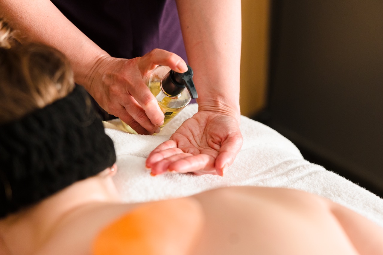 A person applies massage oil onto their hand above the back of a reclining person.