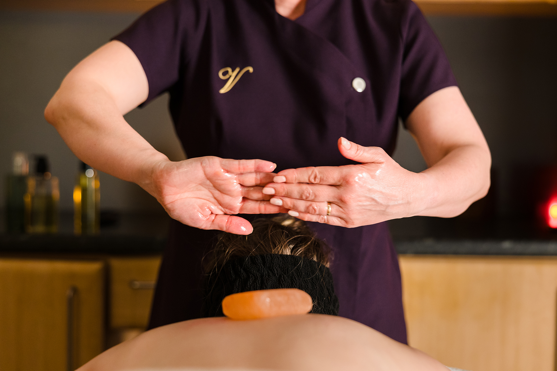 A person in a spa uniform practices reiki over the back of a client with a hot stone placed on the client's back.