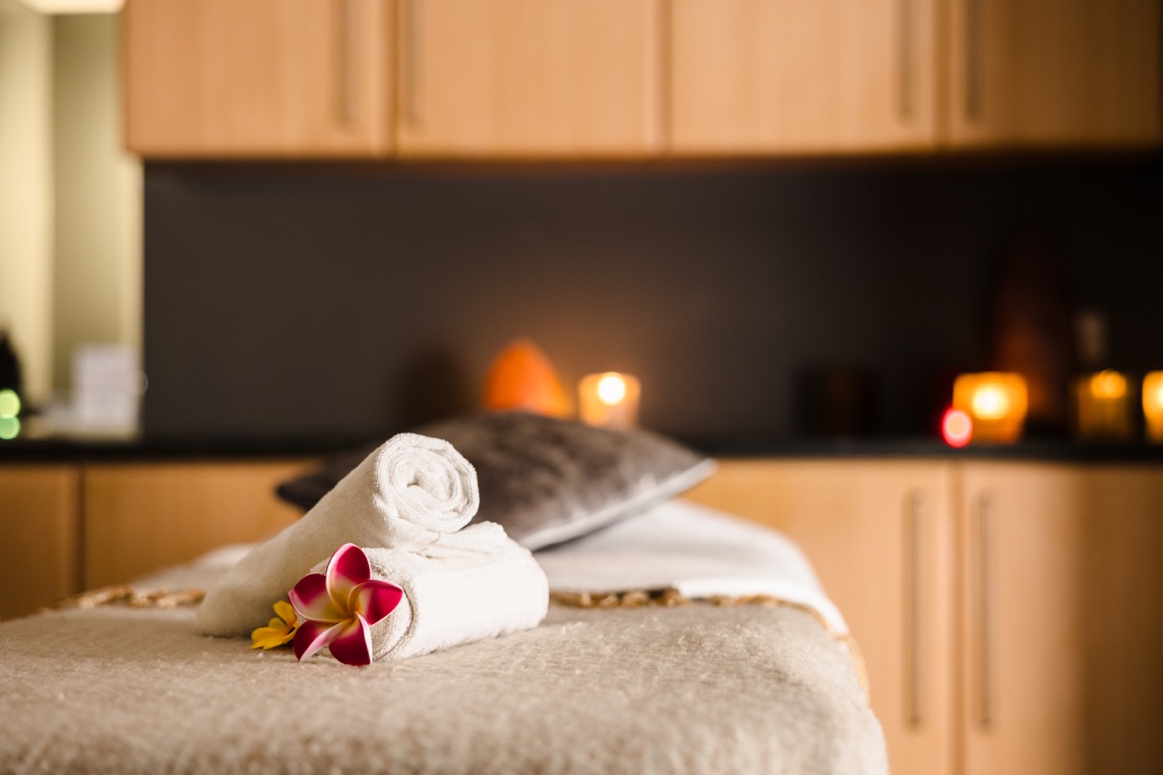 A massage table with rolled towels and plumeria flowers in a dimly lit room, candles glowing softly in the background.