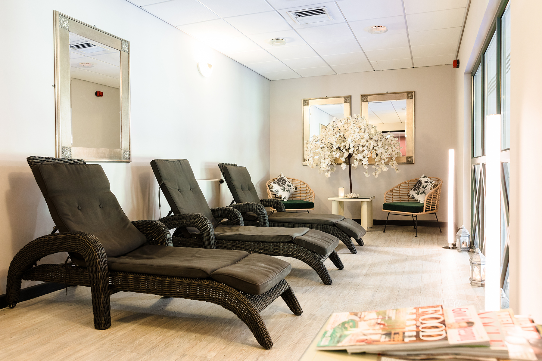 A relaxing lounge area with four wicker chaise lounges, two armchairs, decorative mirrors, and a flower display. Magazines are placed on a table in the foreground.