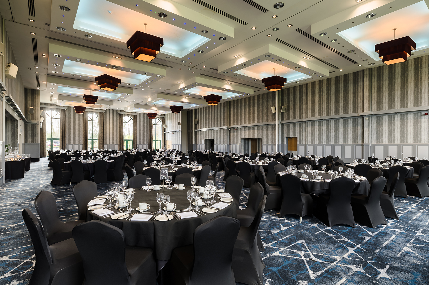 A banquet hall set up with round tables, black chairs, and elegant place settings, illuminated by modern ceiling lights.