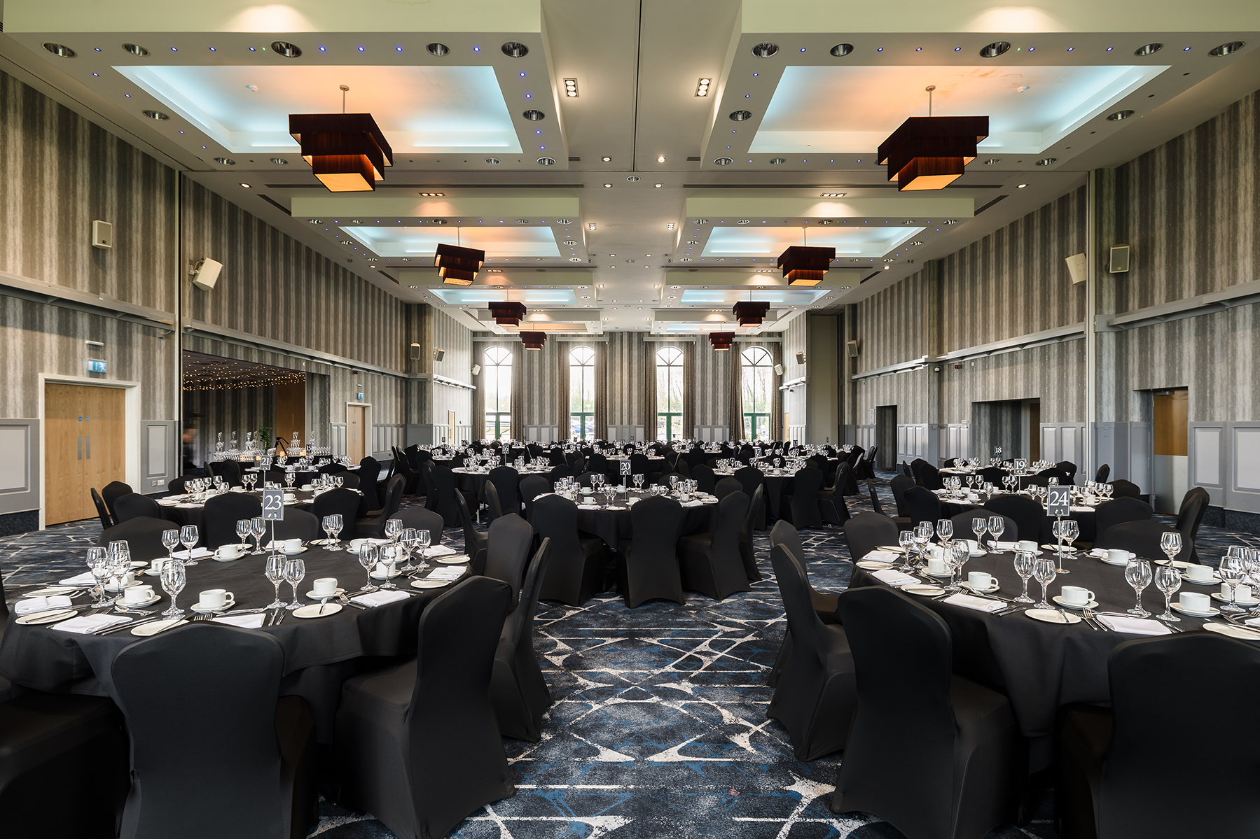 A banquet hall with round tables covered in black tablecloths, set with white plates, glasses, and silverware, chairs draped in black covers, and chandeliers on the ceiling.