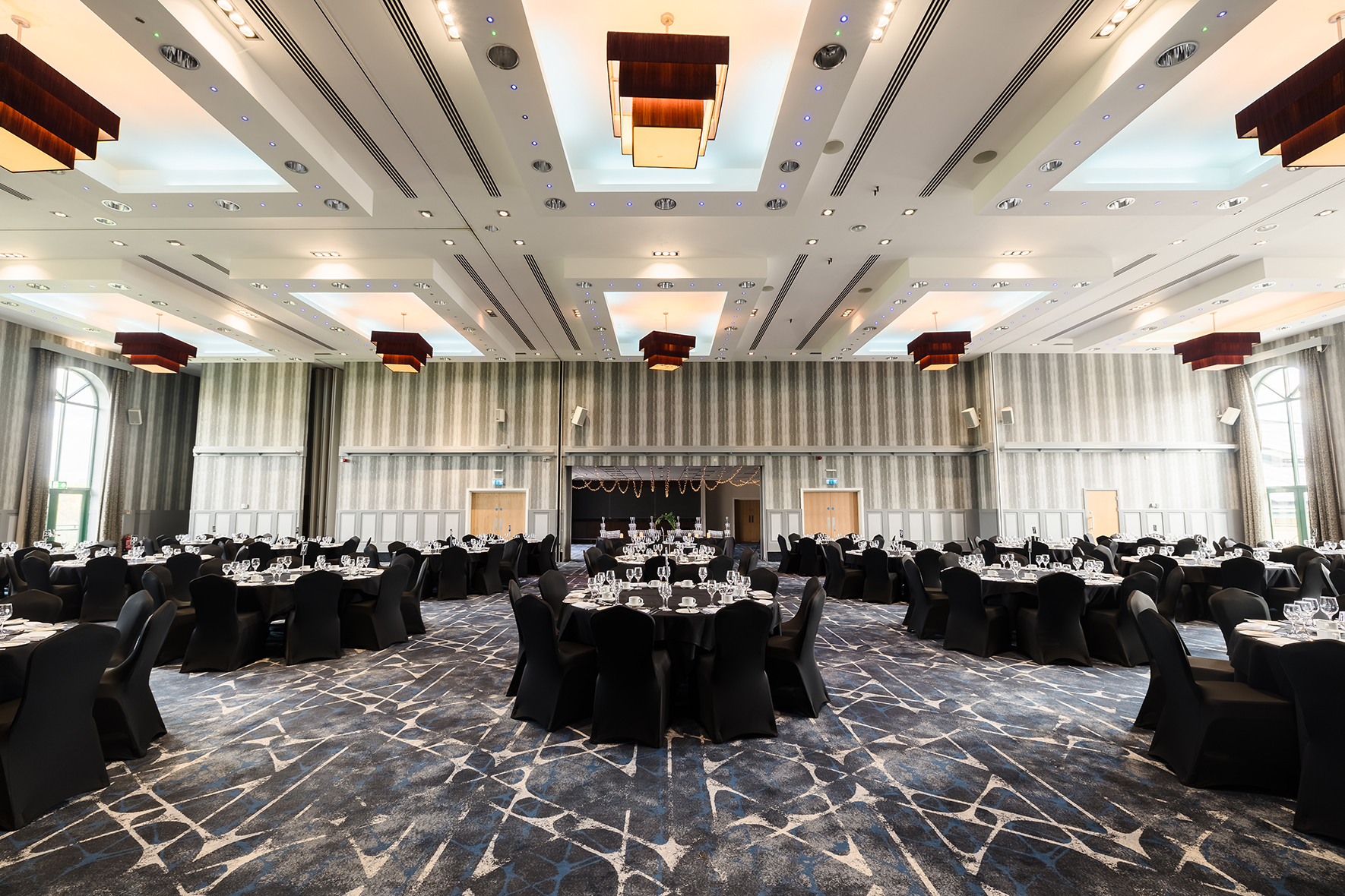 Spacious banquet hall with elegantly set round tables, black chair covers, and patterned carpet. The ceiling has modern light fixtures and large windows on the sides.