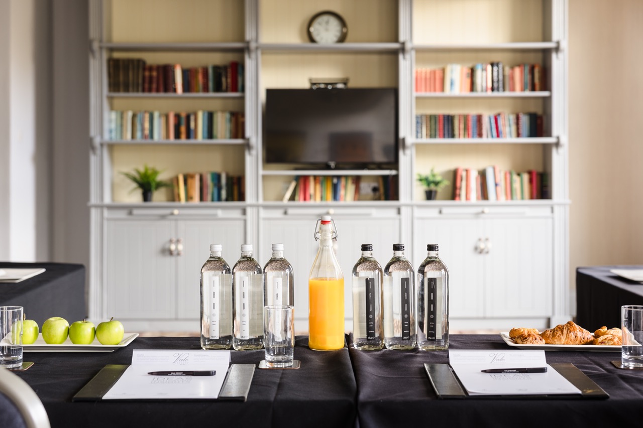 Conference room setup with bottled water, a glass bottle of orange juice, apples, and croissants on a table. A bookshelf and television are in the background.