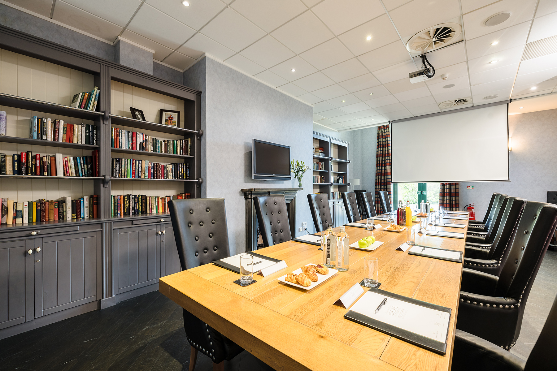 A conference room with a long wooden table, black chairs, a white screen, shelves with books, and a wall-mounted TV.