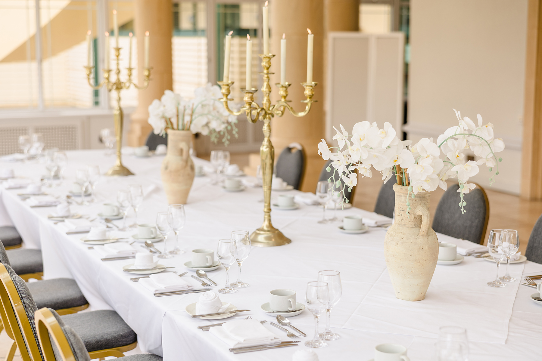 A long, elegantly set table with white cloth, chairs, tall golden candelabras, white flowers in vases, and neatly arranged dinnerware and glassware.