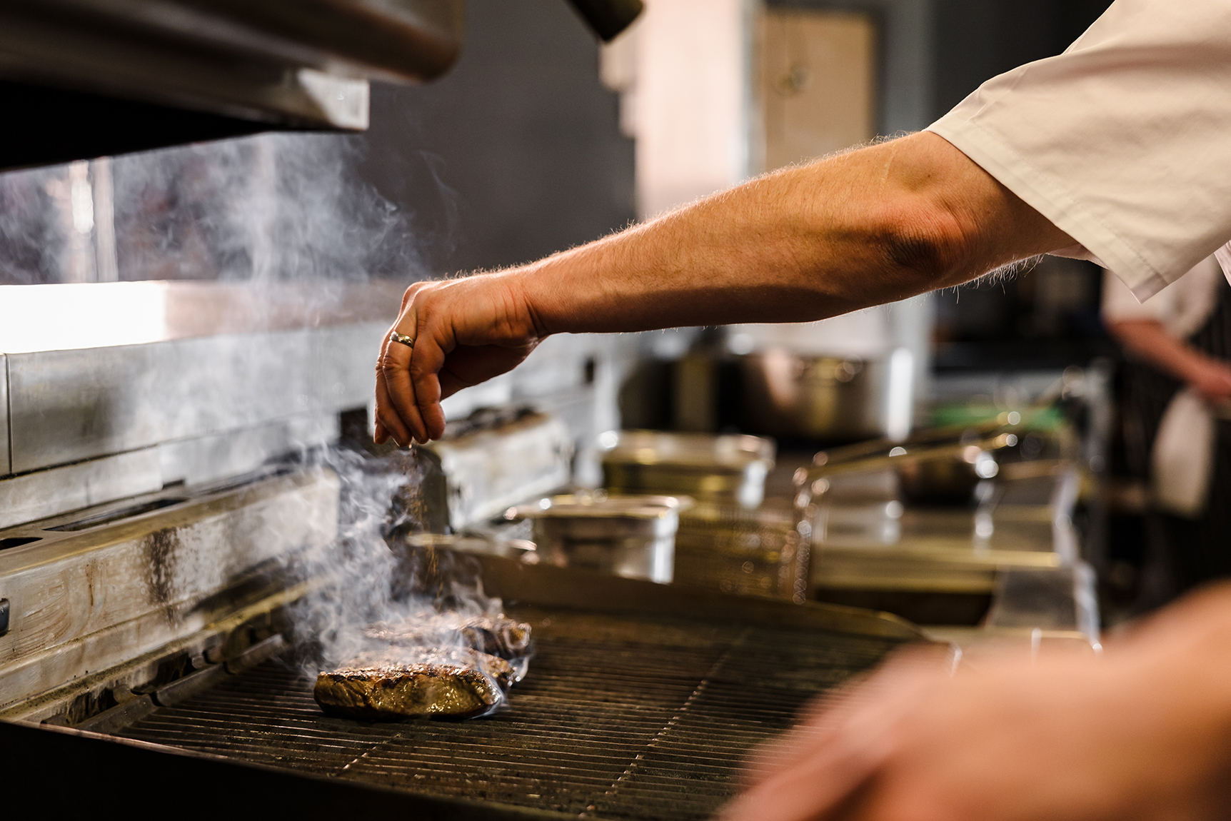 Chef seasoning a steak on a grill with steam rising, in a professional kitchen setting.