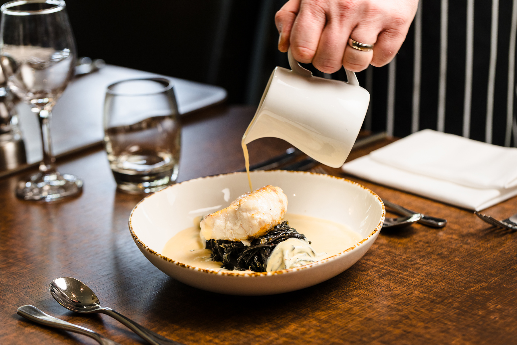 A person pours sauce from a white pitcher over a plated dish on a table set with utensils and glasses.