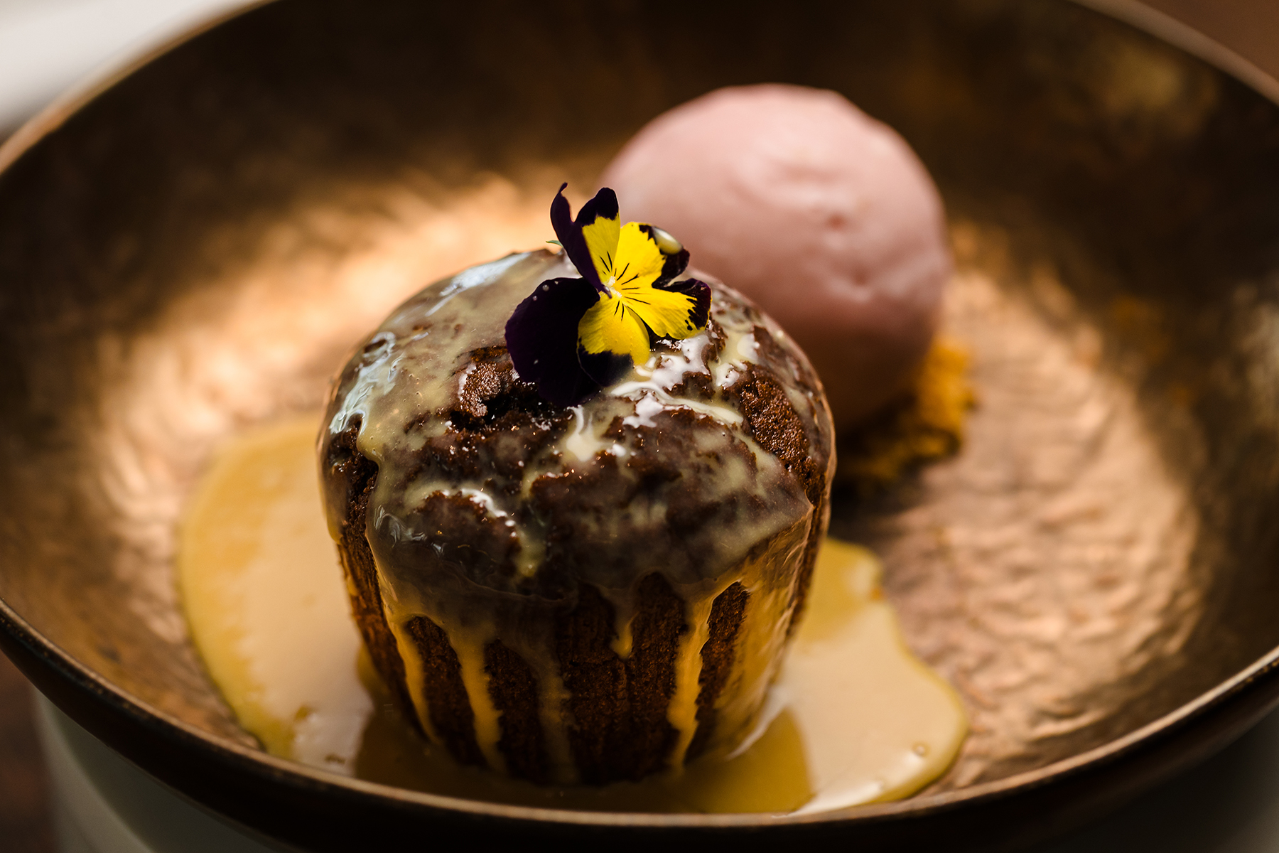 A chocolate muffin with glaze and a flower on top, served with a scoop of pink ice cream in a rustic bowl.