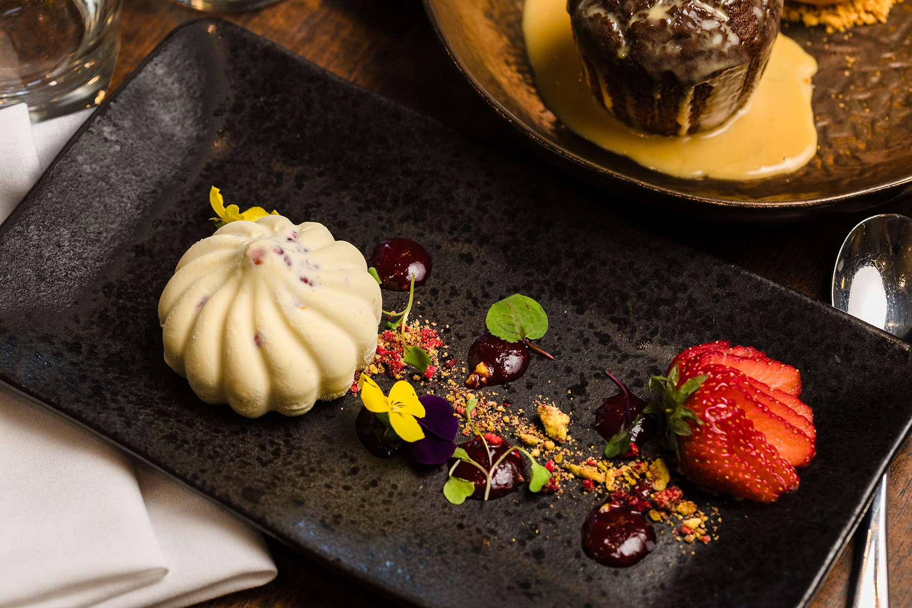 A dessert plate with a white mousse cake, garnished with edible flowers, strawberries, and fruit sauce dots, next to a napkin and another dessert in the background.