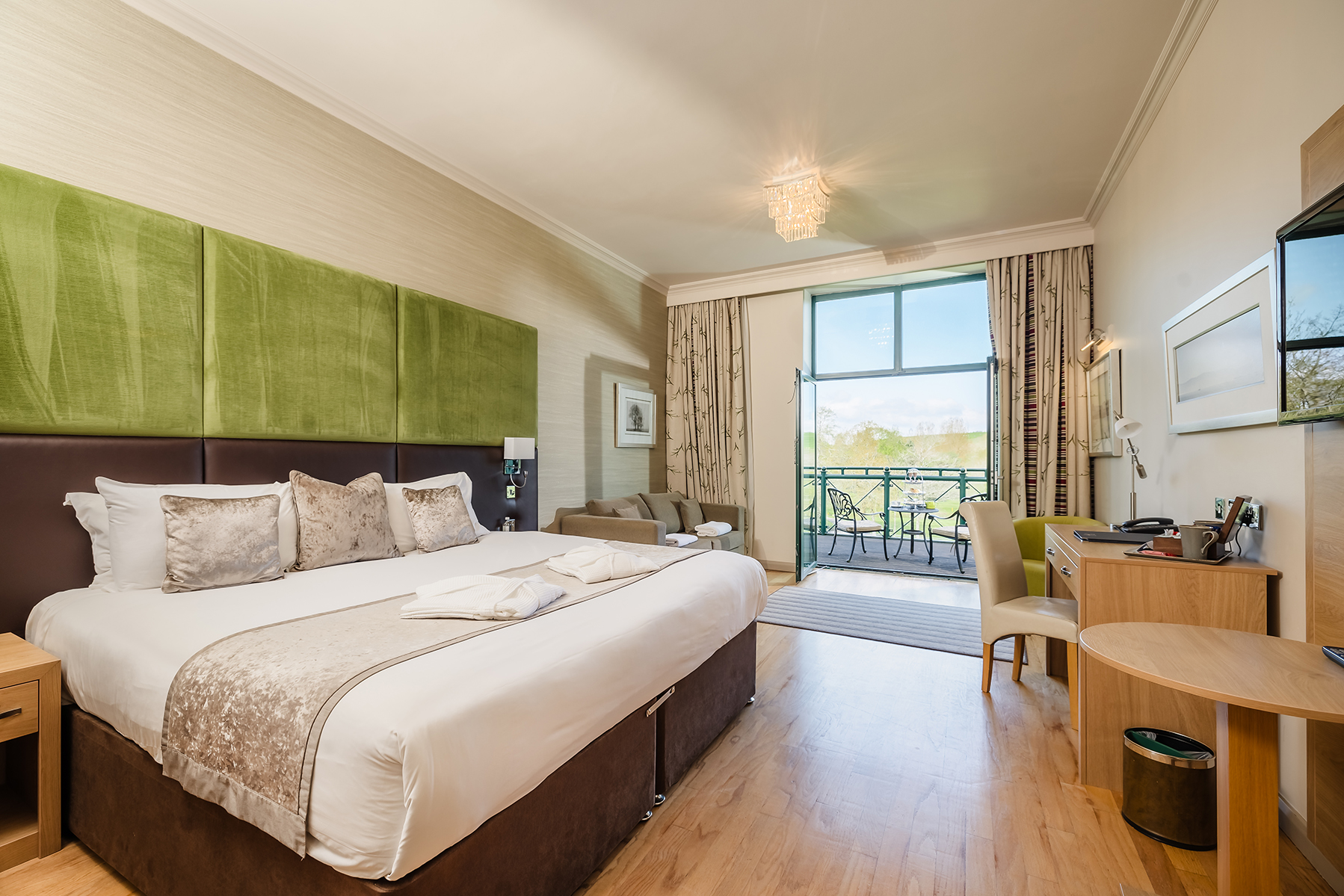 A hotel room with a large bed, green headboard, small desk, chair, and TV. Balcony with outdoor seating is visible through glass door. Bright and neatly organized.