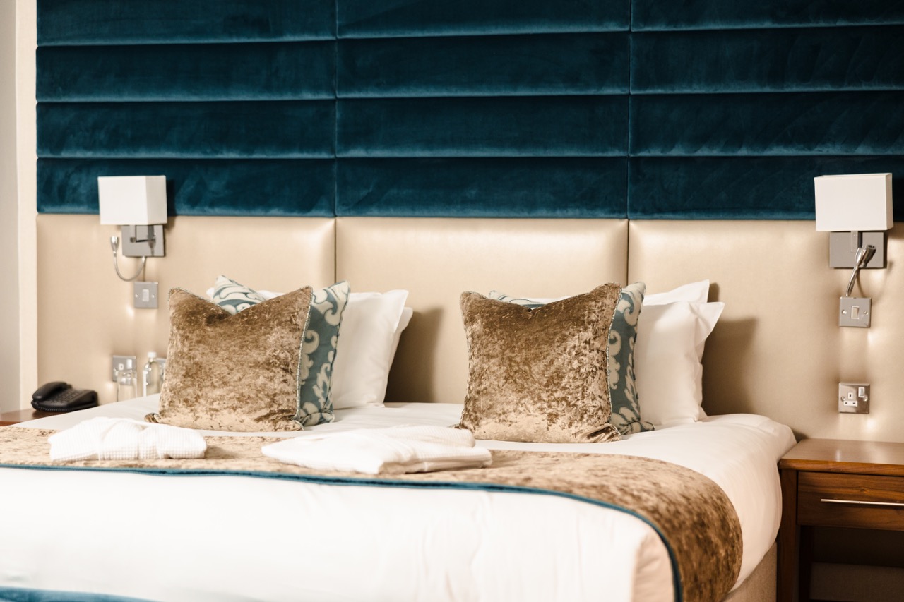 A neatly made bed with plush pillows and a folded throw blanket. Two wall-mounted lamps and a padded teal headboard are above the bed.