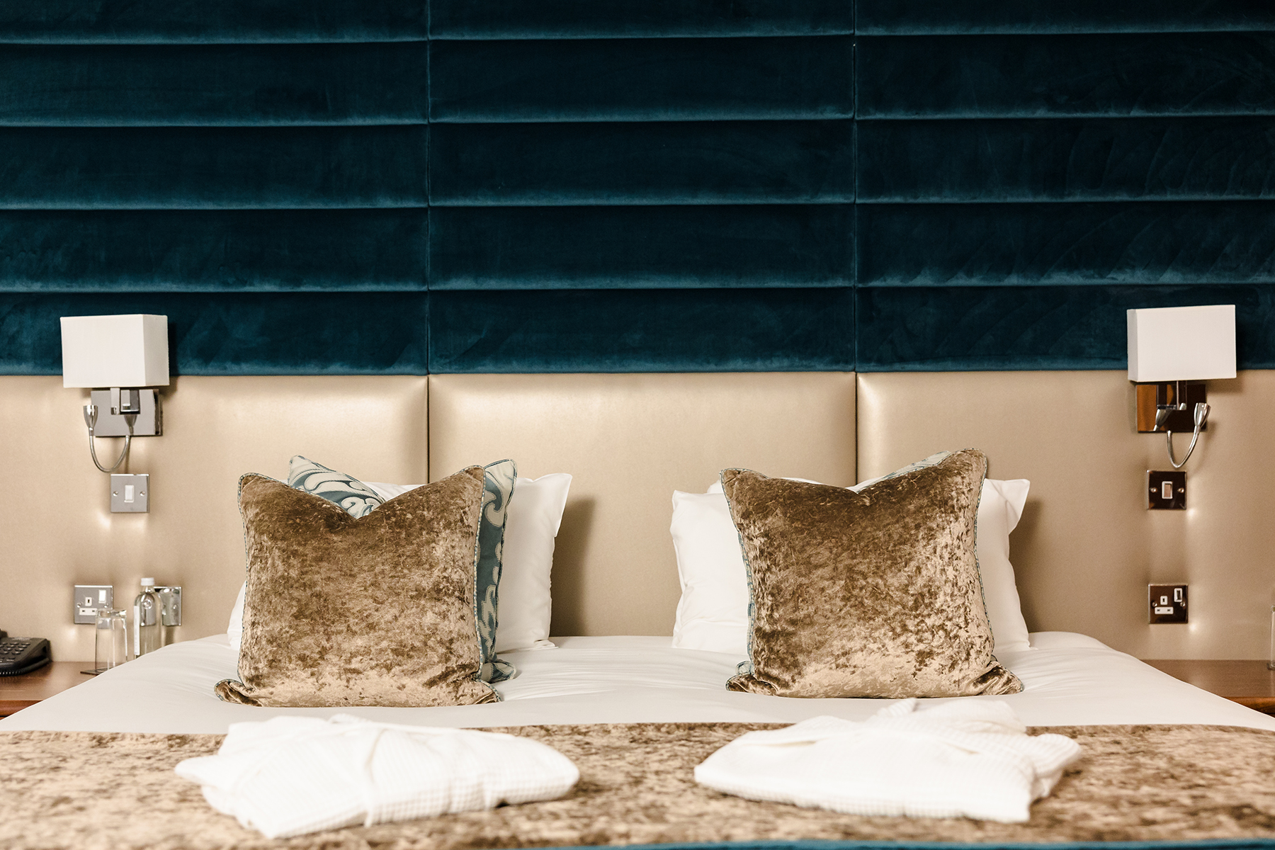 A hotel room bed with two plush pillows and a textured headboard. There are wall-mounted lamps on either side and two folded robes at the foot of the bed.