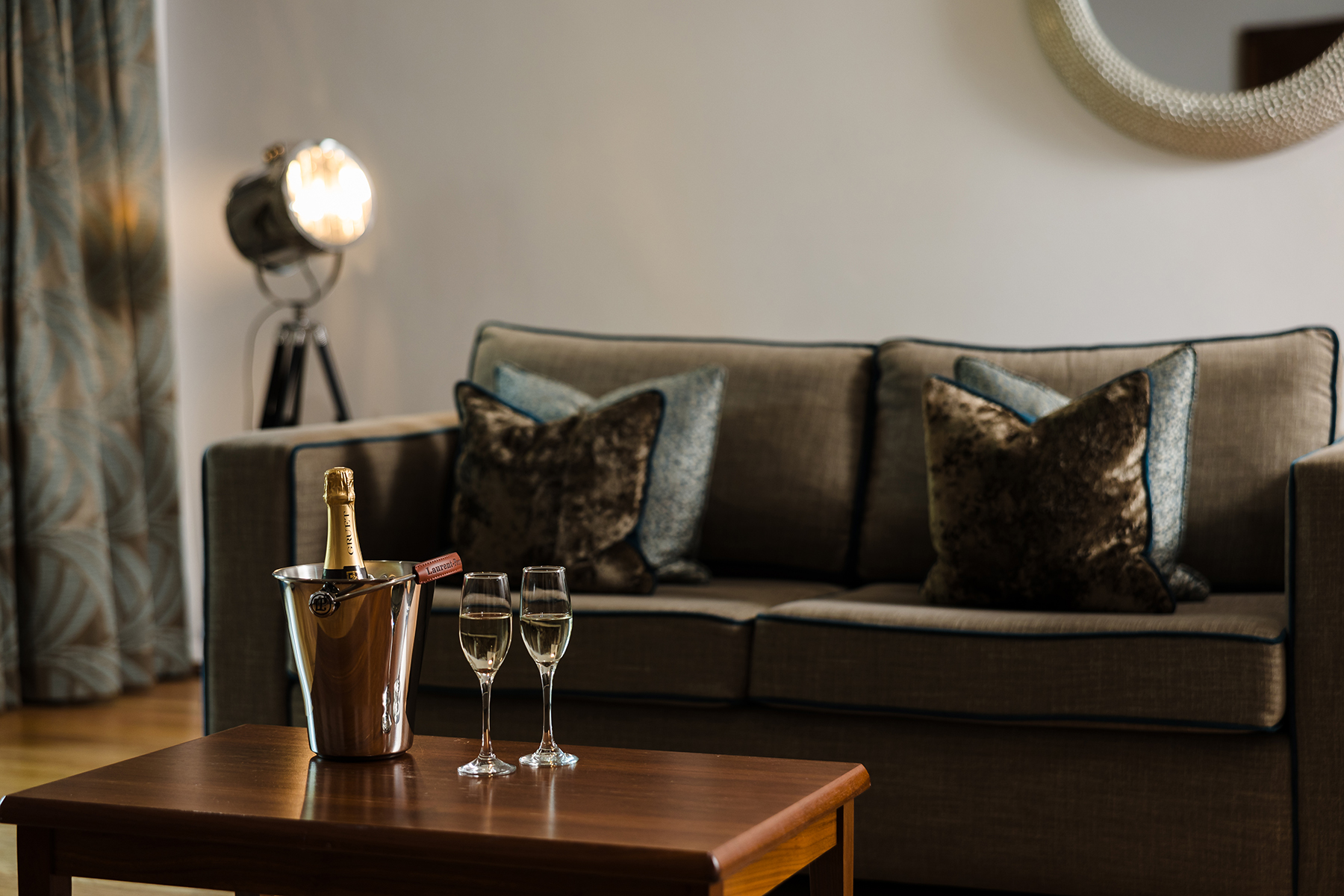 A cozy living room features a gray sofa with dark pillows, a wooden table with a champagne bottle in an ice bucket and two glasses, and a spotlight lamp in the background.