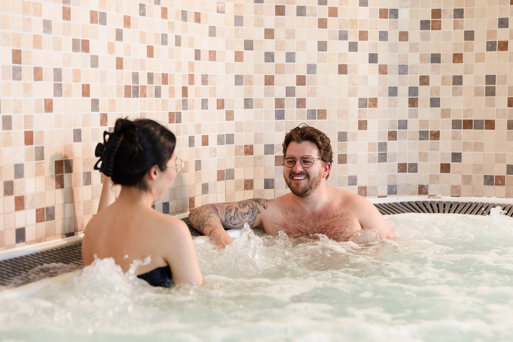 Two people relaxing in a hot tub with mosaic tile walls in the background.