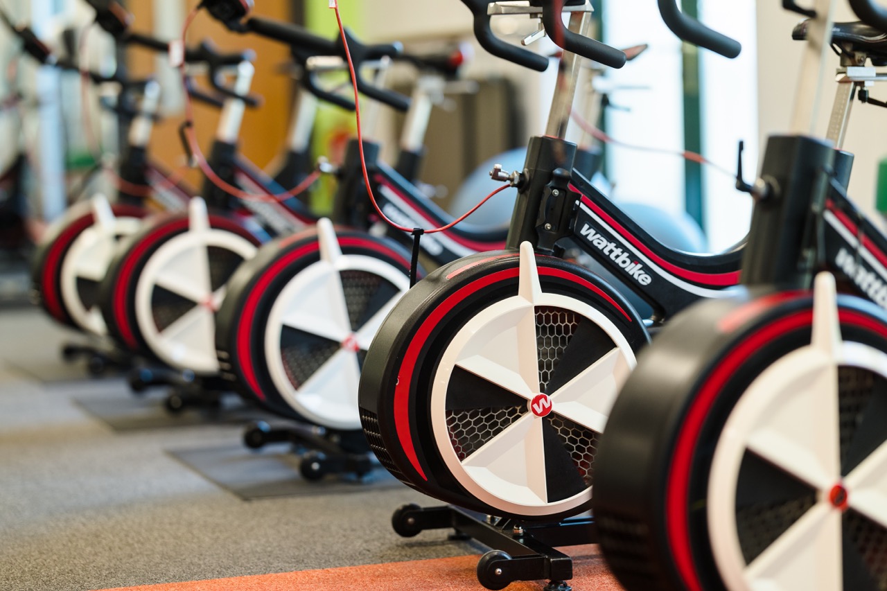 A row of stationary exercise bikes in a gym, each with a large fan wheel and digital displays.