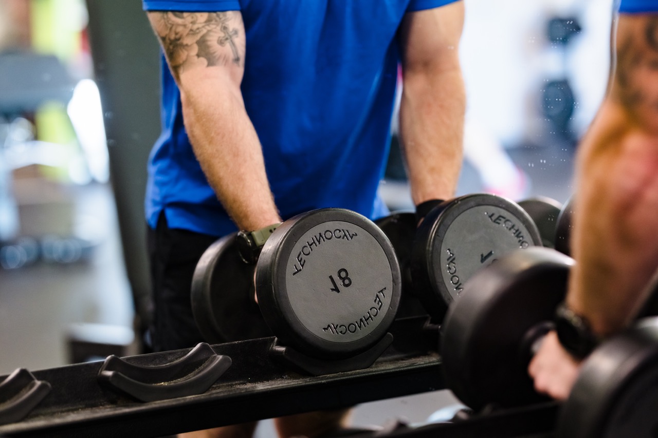 Person in a blue shirt lifting a pair of dumbbells in front of a mirror at the gym.