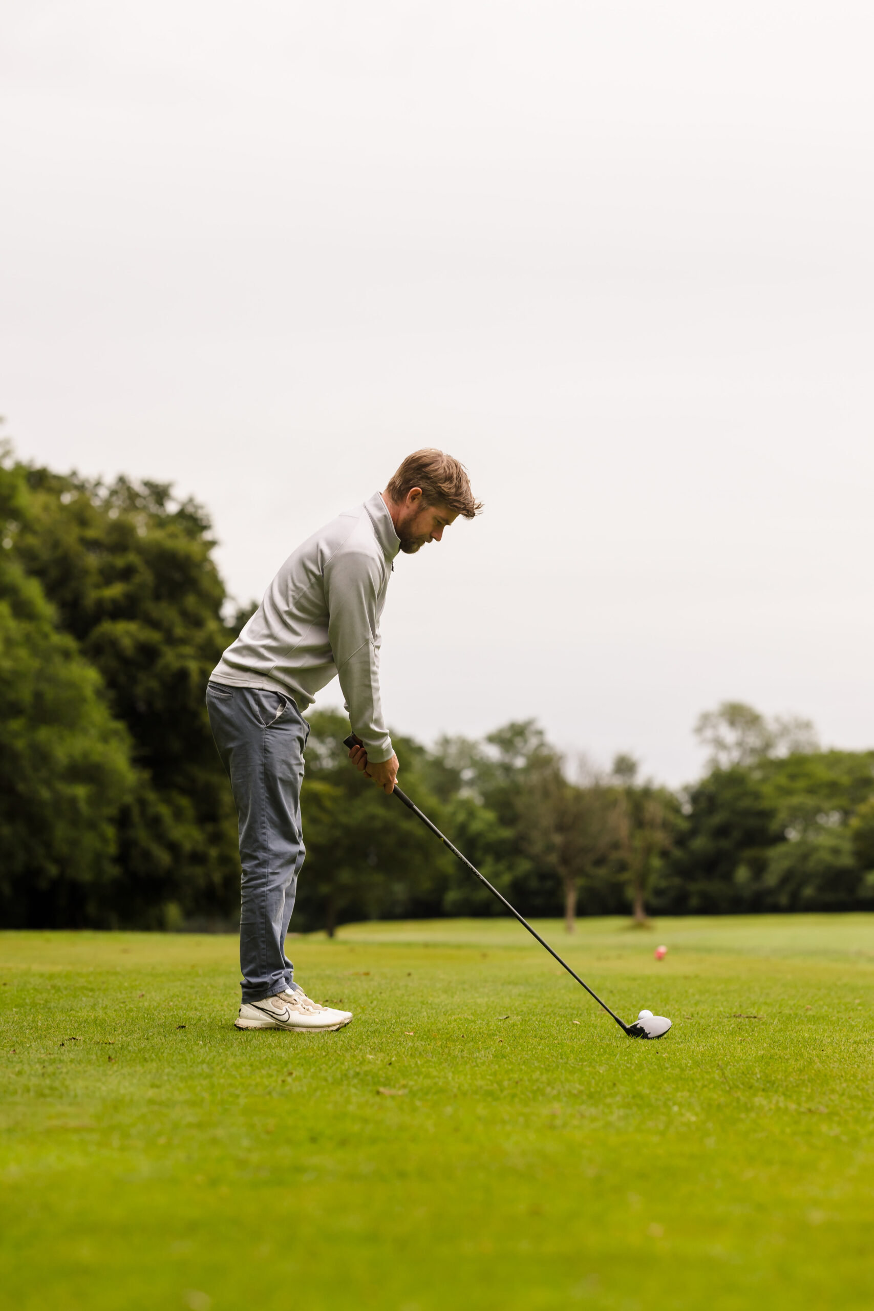 A man in a white sweater and gray pants prepares to hit a golf ball on a lush green golf course. Trees are visible in the background.