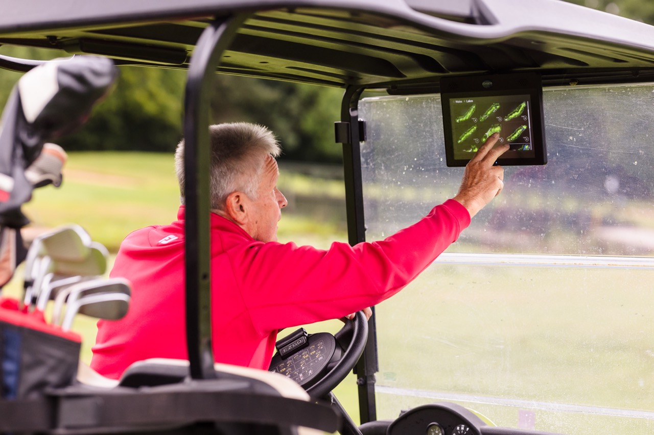 A man in a red jacket interacts with a touchscreen in a golf cart on a golf course. Golf clubs are visible in the back.