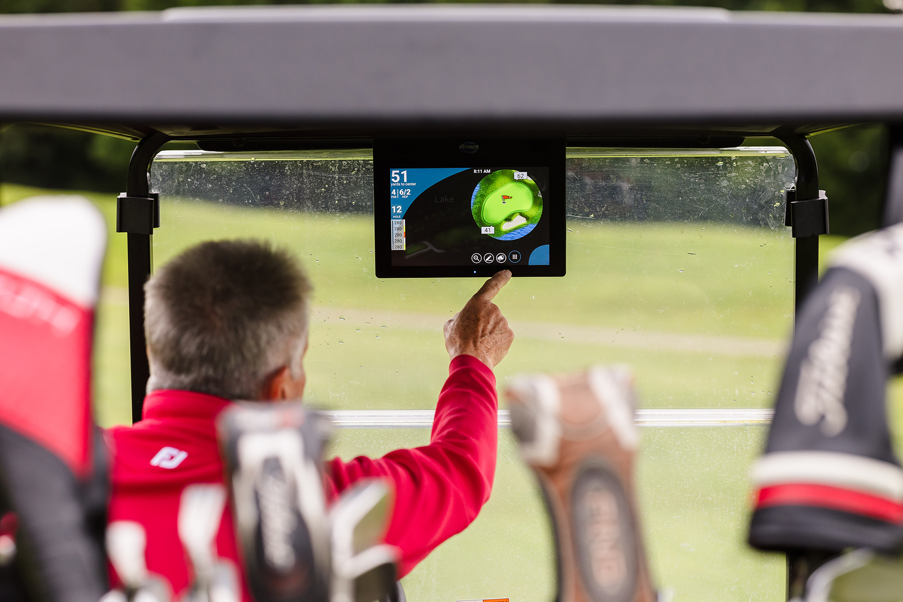 A person in a red jacket operates a touchscreen golf GPS device inside a golf cart.