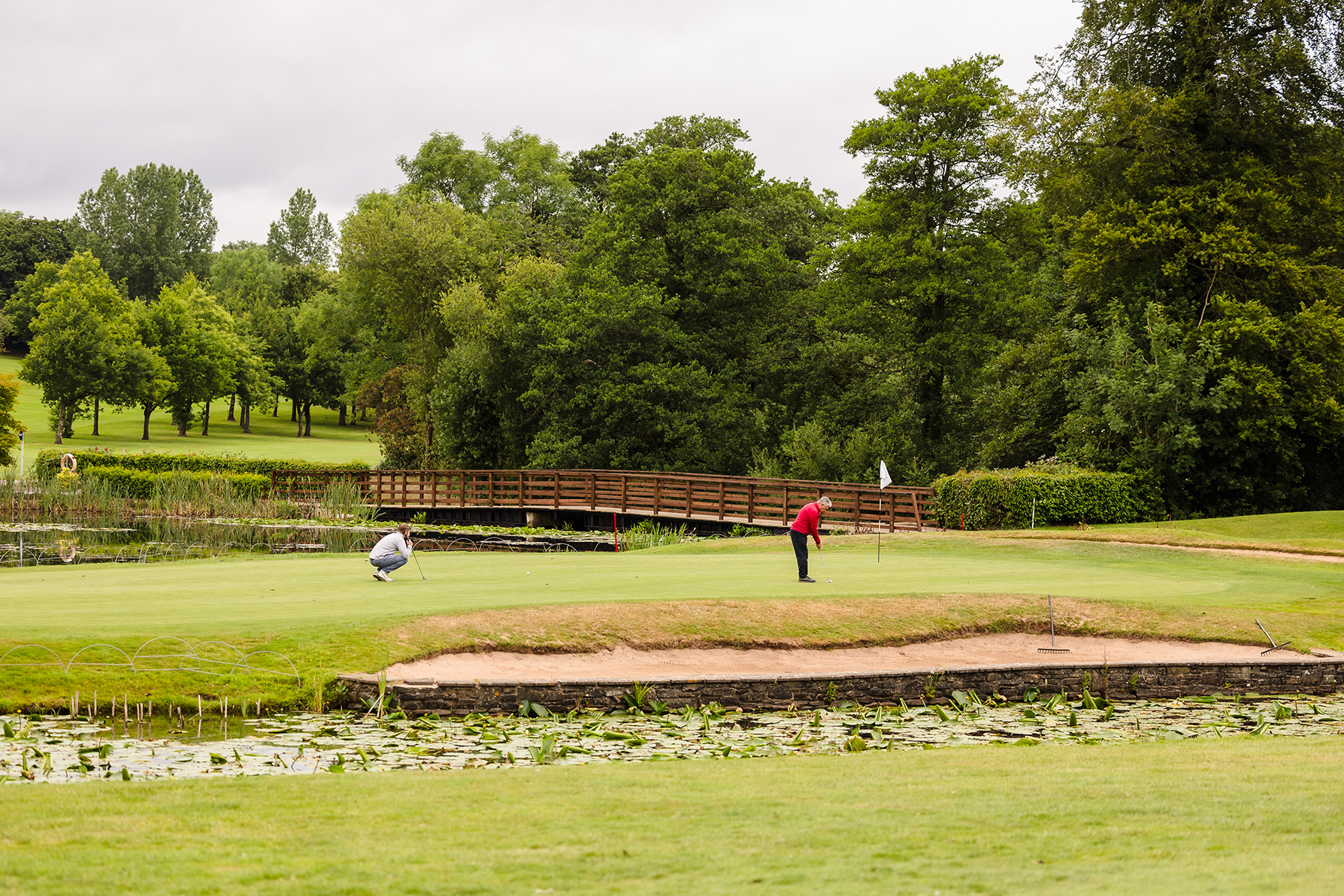 Two people on a golf green, one putting while the other observes. A wooden bridge and trees are in the background.