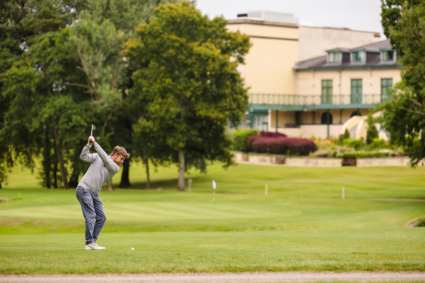 A person is playing golf on a green course, preparing to swing. Trees and a large building are visible in the background.