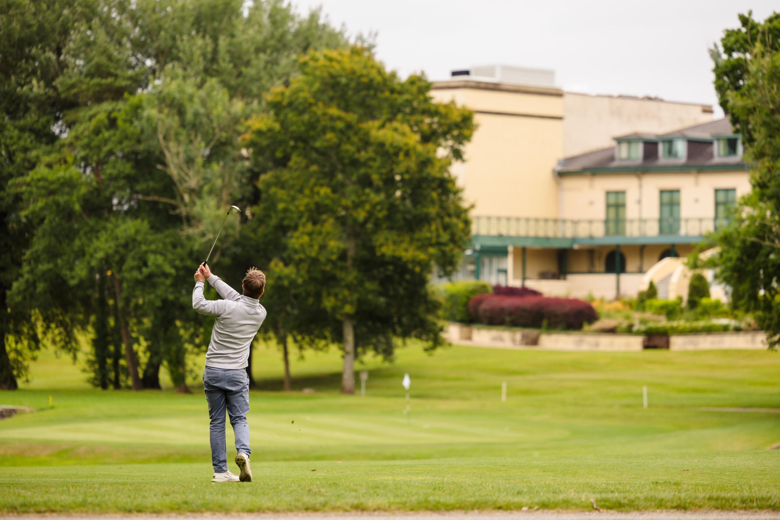 A person swings a golf club on a green golf course with trees and a large building in the background.