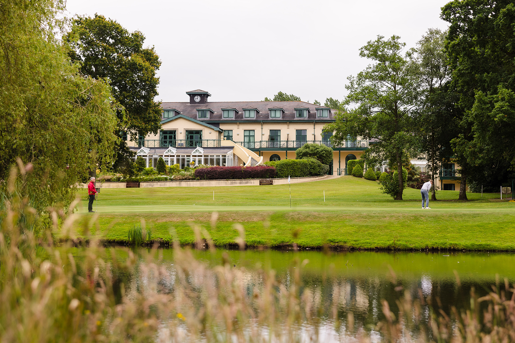 Two people on a manicured lawn in front of a large building, surrounded by trees, with a pond in the foreground.