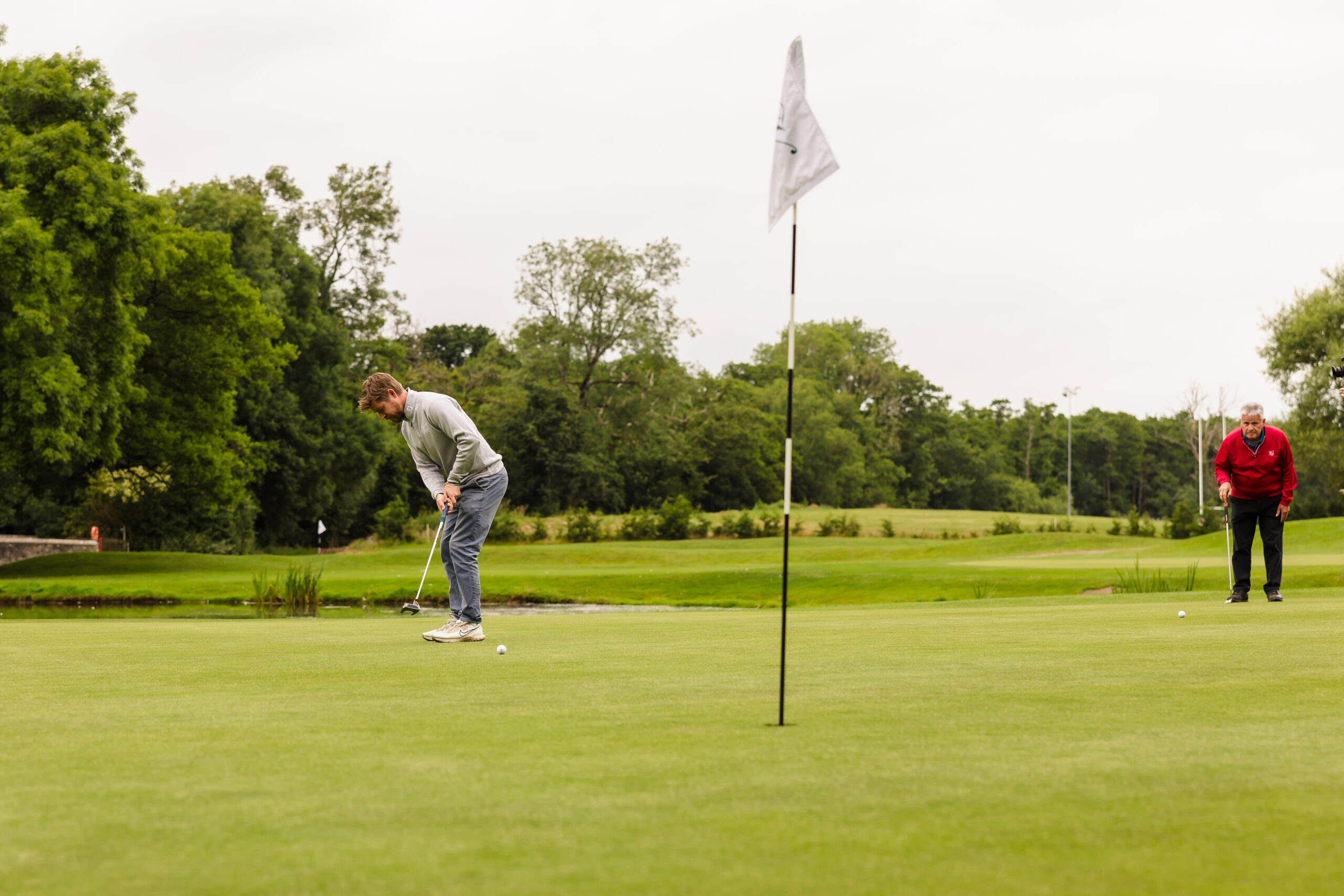 A person wearing a gray sweater putts on a golf course green while another person in a red sweater watches. A flagstick is visible in the foreground.