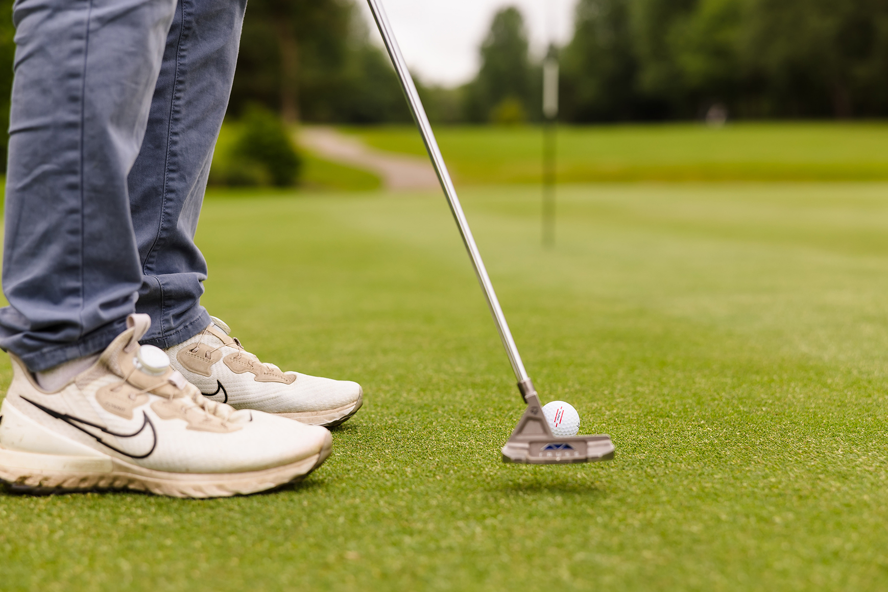 A person in jeans and white sneakers is about to putt a golf ball on a green.