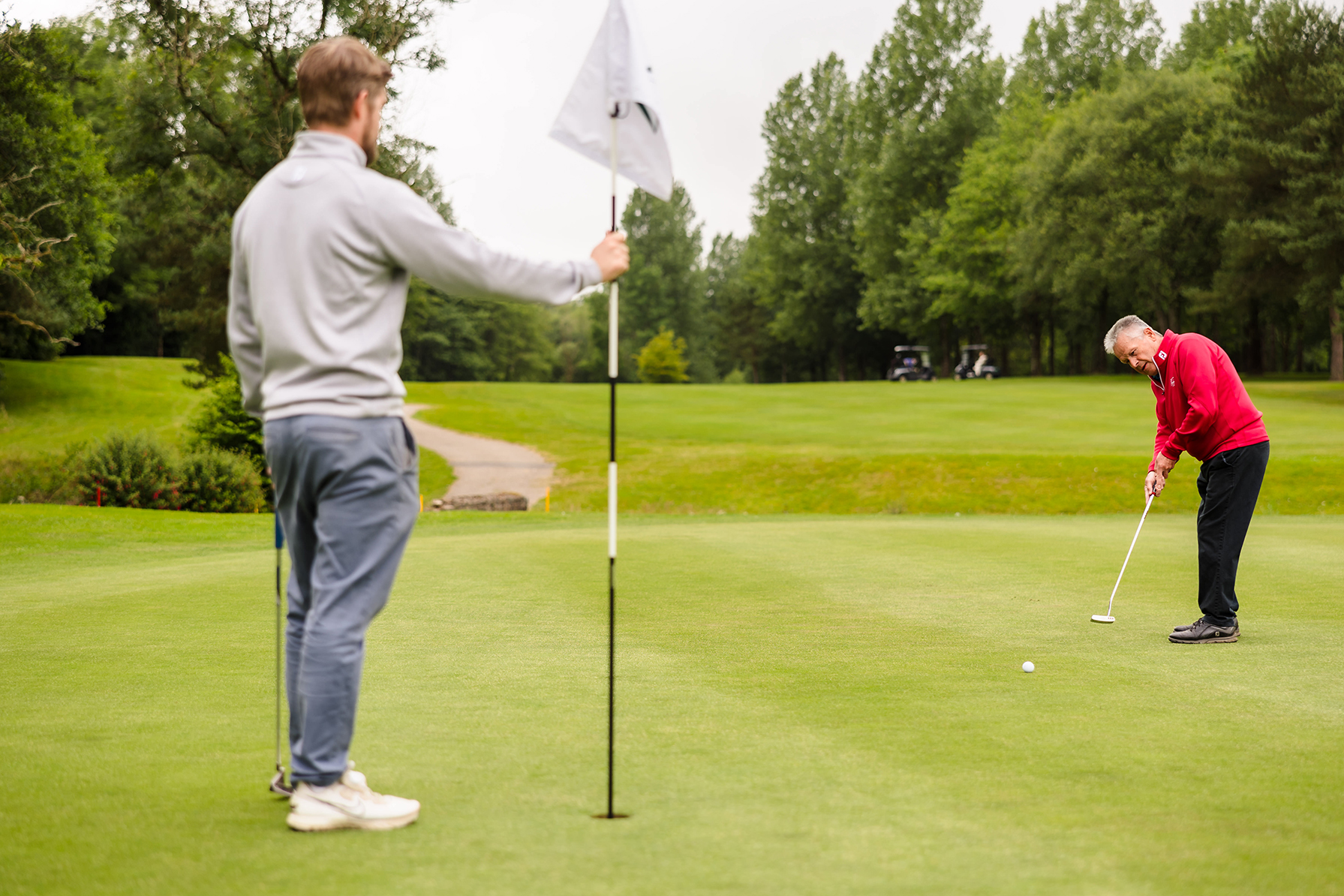 Two people are on a golf course. One person in a red sweater is putting, while the other in a gray jacket holds the flagstick. Greenery surrounds the scene.