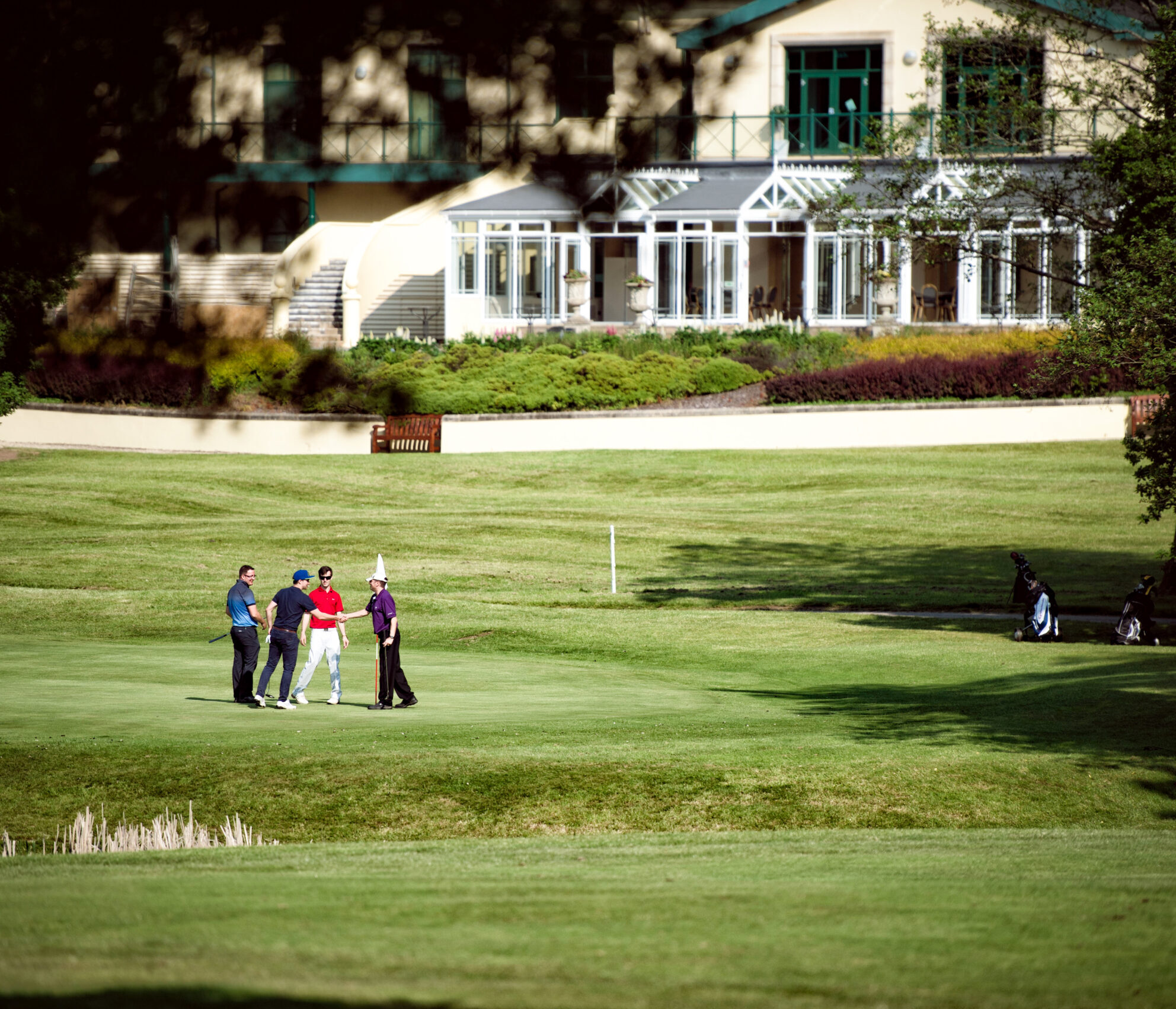 Four people stand on a golf course, shaking hands near a clubhouse in the background. Golf bags are visible on the grass.