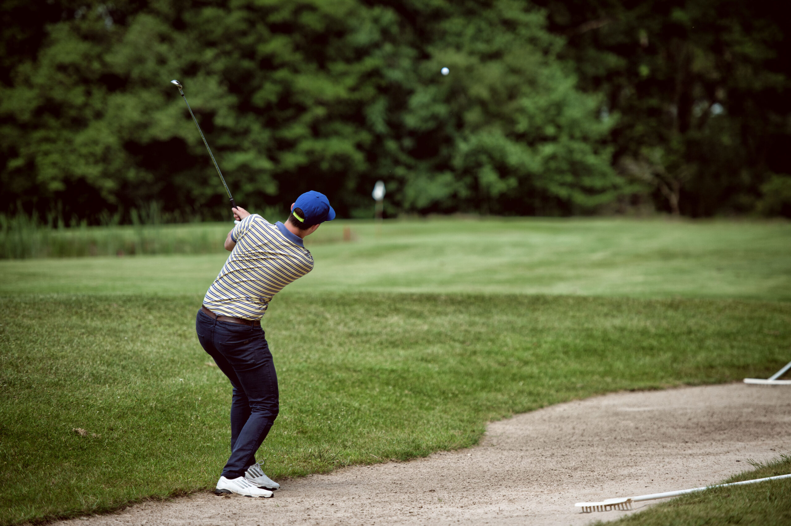 A person in a striped shirt and blue cap is swinging a golf club on a lush green golf course near a sand trap.