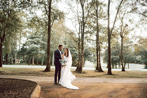 A bride and groom stand together on a path in a park, surrounded by tall trees.