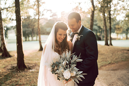 A bride and groom stand in a sunlit park. The bride holds a bouquet and wears a veil. Both are smiling. Trees and a path are visible in the background.