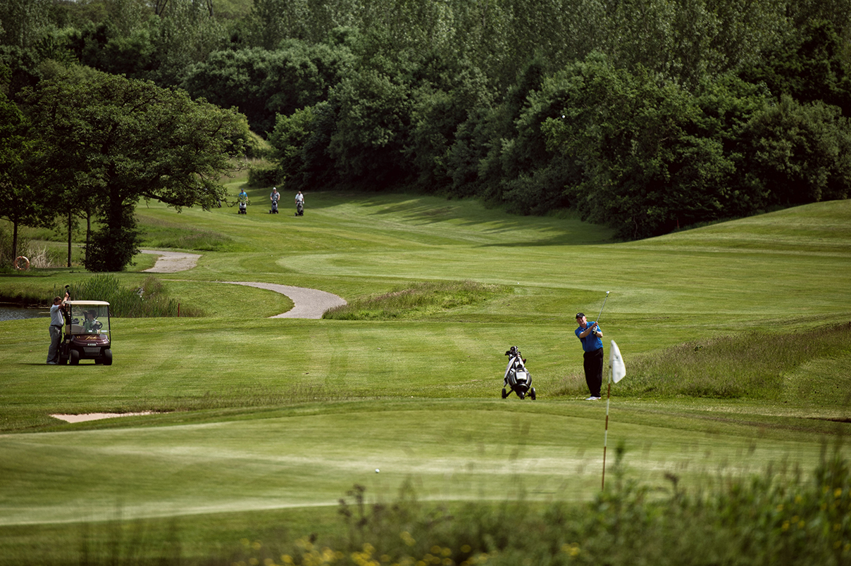 Golfer on a lush green course, preparing to swing near a flag, with a golf cart and trees in the background.