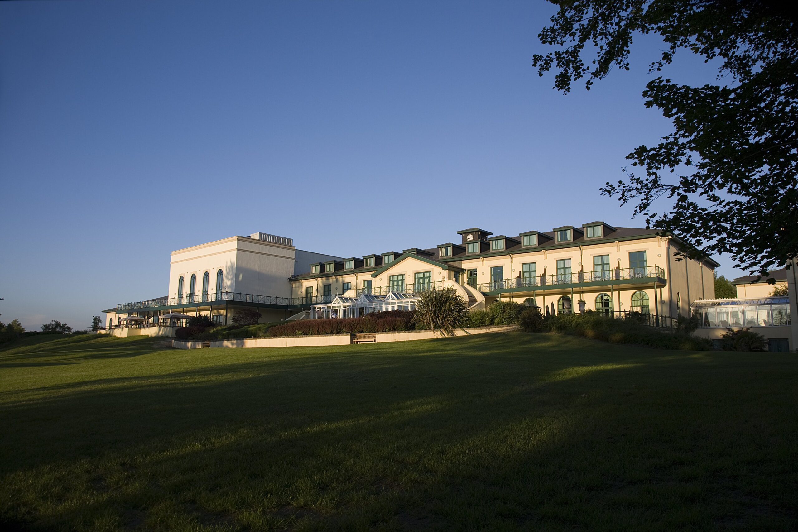 A large hotel building with green lawns, arched windows, and a modern section. It's a clear day with a blue sky.