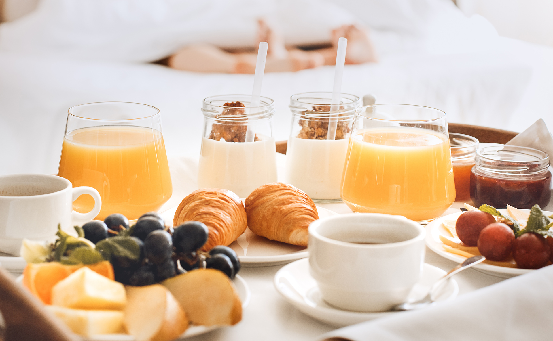 Breakfast tray with croissants, orange juice, yogurt with granola, coffee, and a plate of grapes and sliced fruit on a bed.