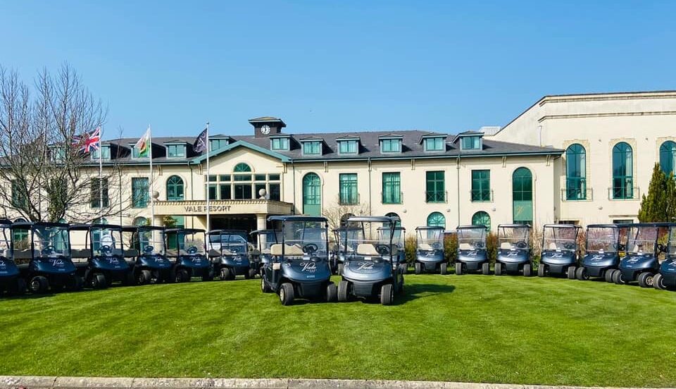 A line of golf carts is parked on a lawn in front of a large building with multiple flags on the roof.