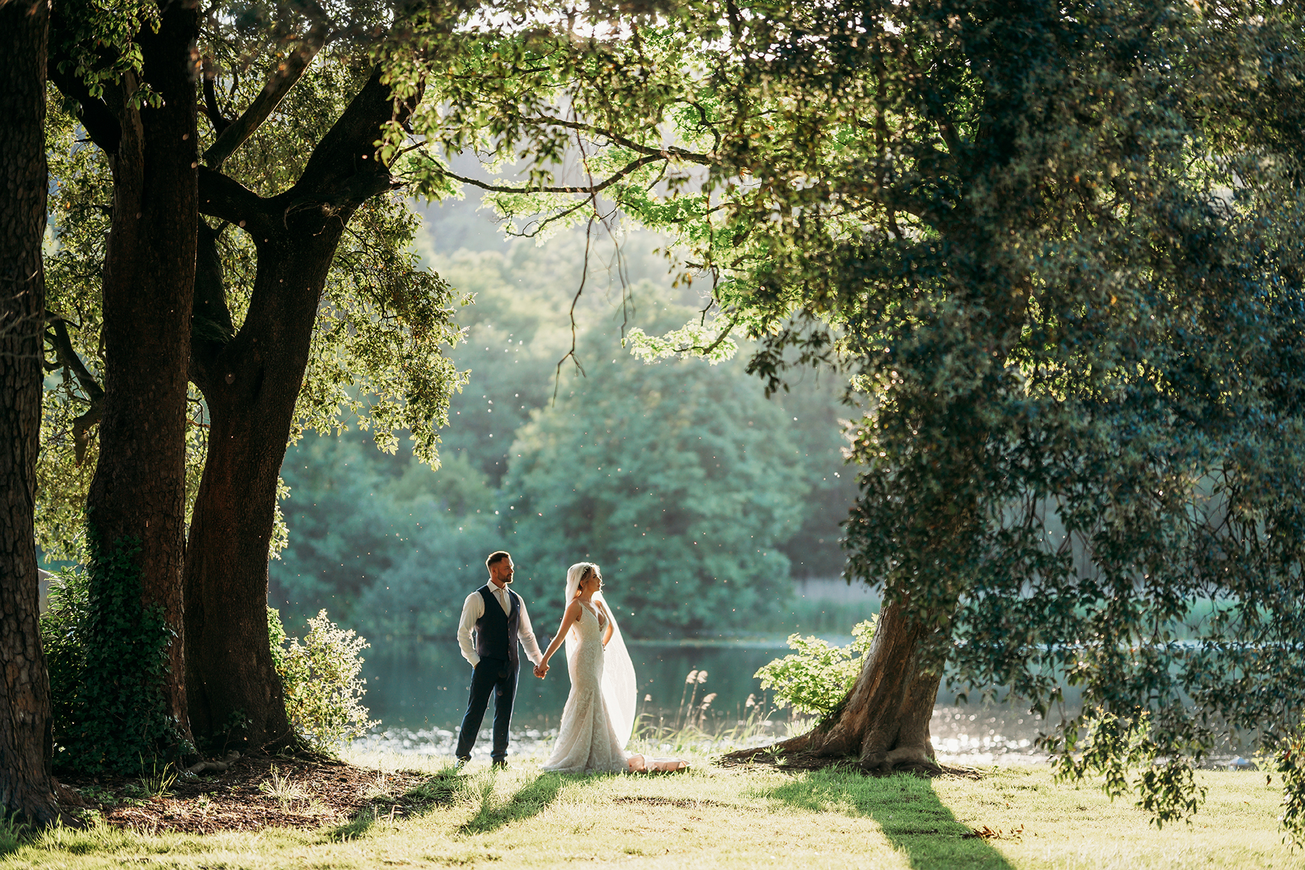 Bride and groom holding hands under trees, standing on a grassy area by a pond, with sunlight filtering through the leaves.