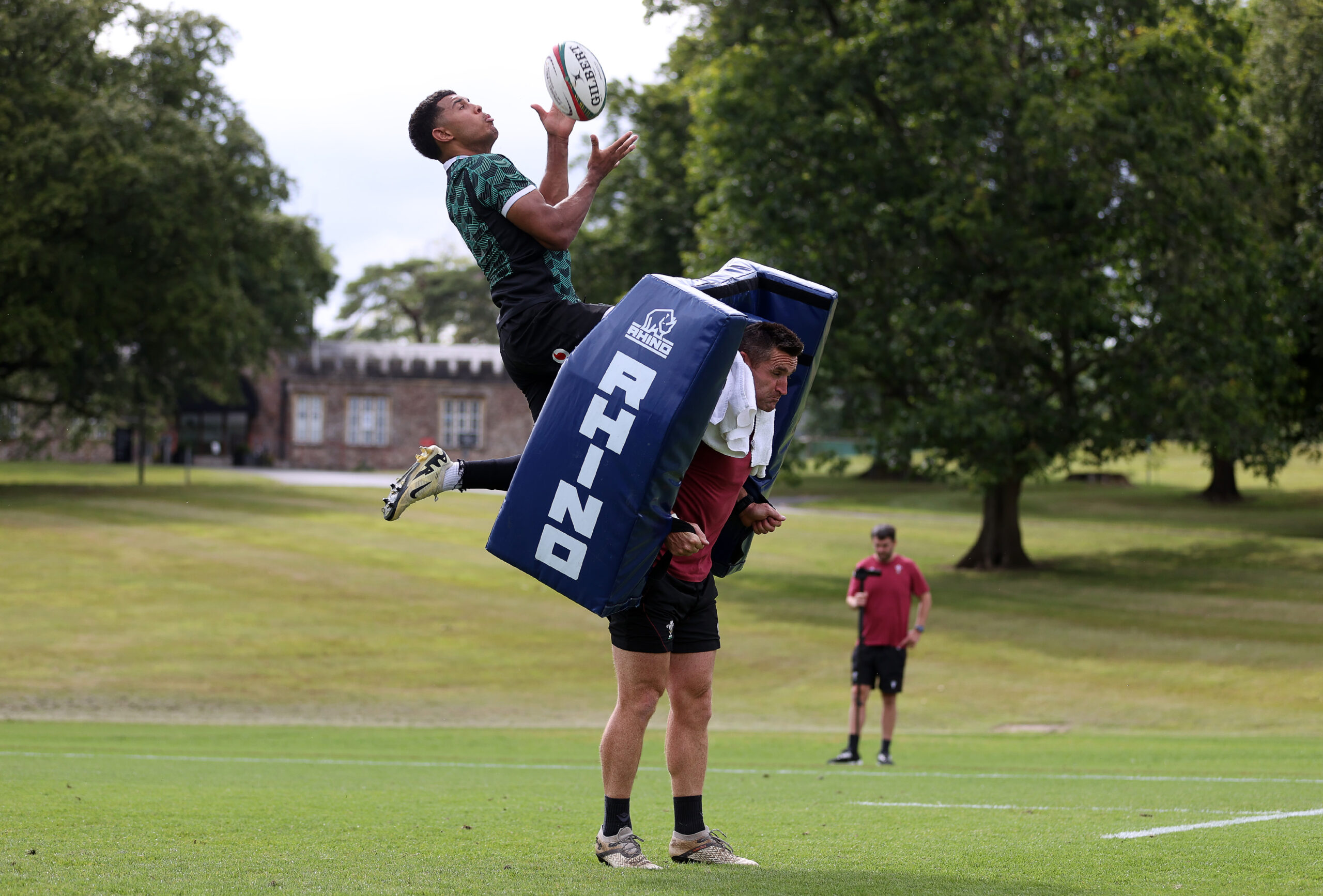 Rugby practice scene: one player catching a ball mid-air, standing on a padded tackle shield held by another player. A coach watches in the background on a grassy field.