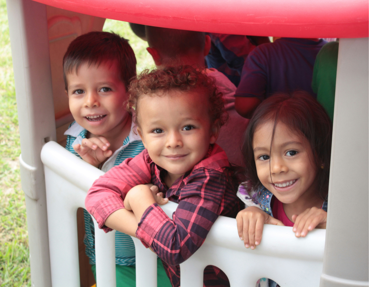 Three children smiling while leaning on a white playhouse fence outdoors.