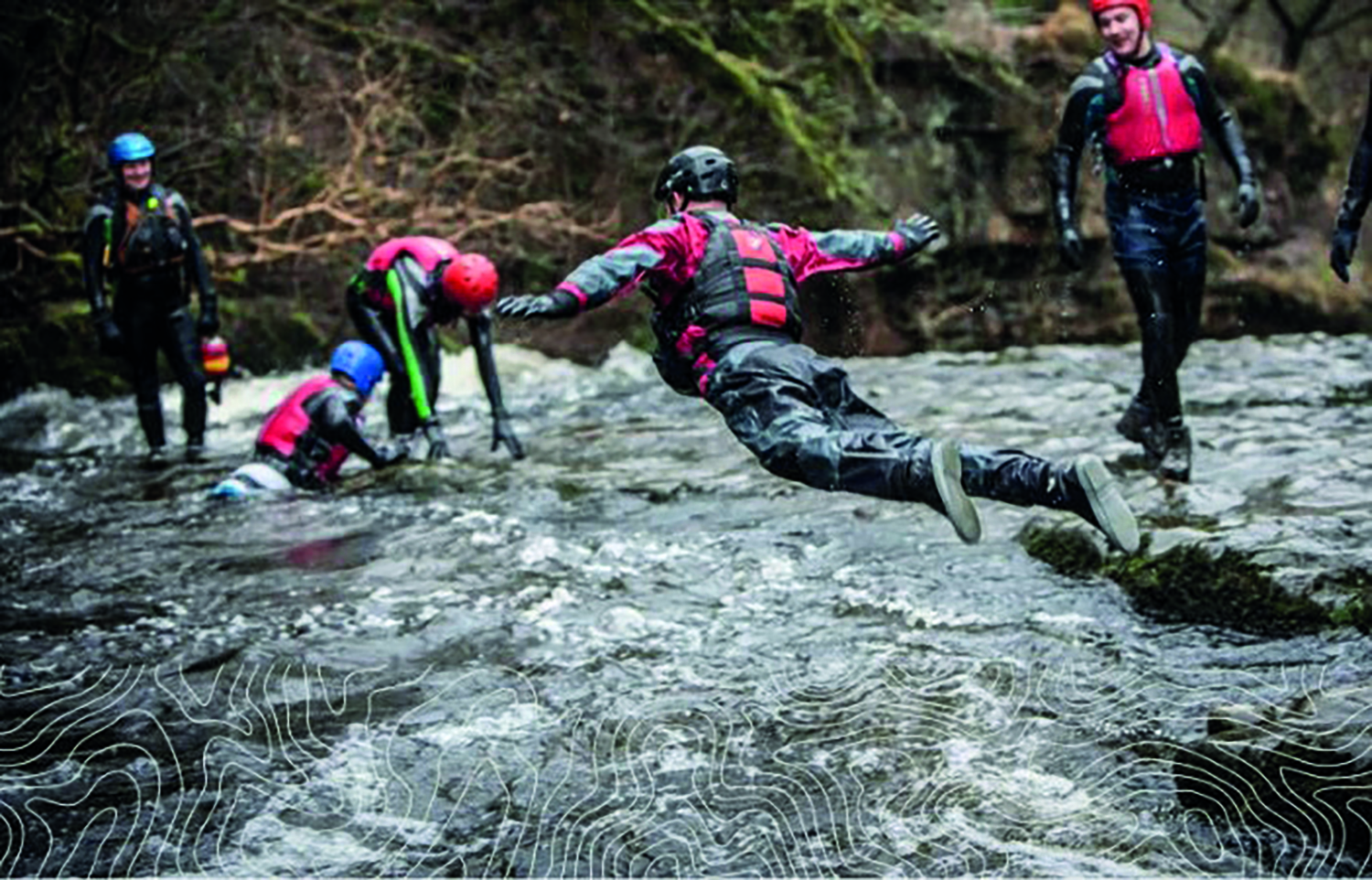 Person in protective gear jumps across a rocky stream while others in similar gear watch, surrounded by trees.