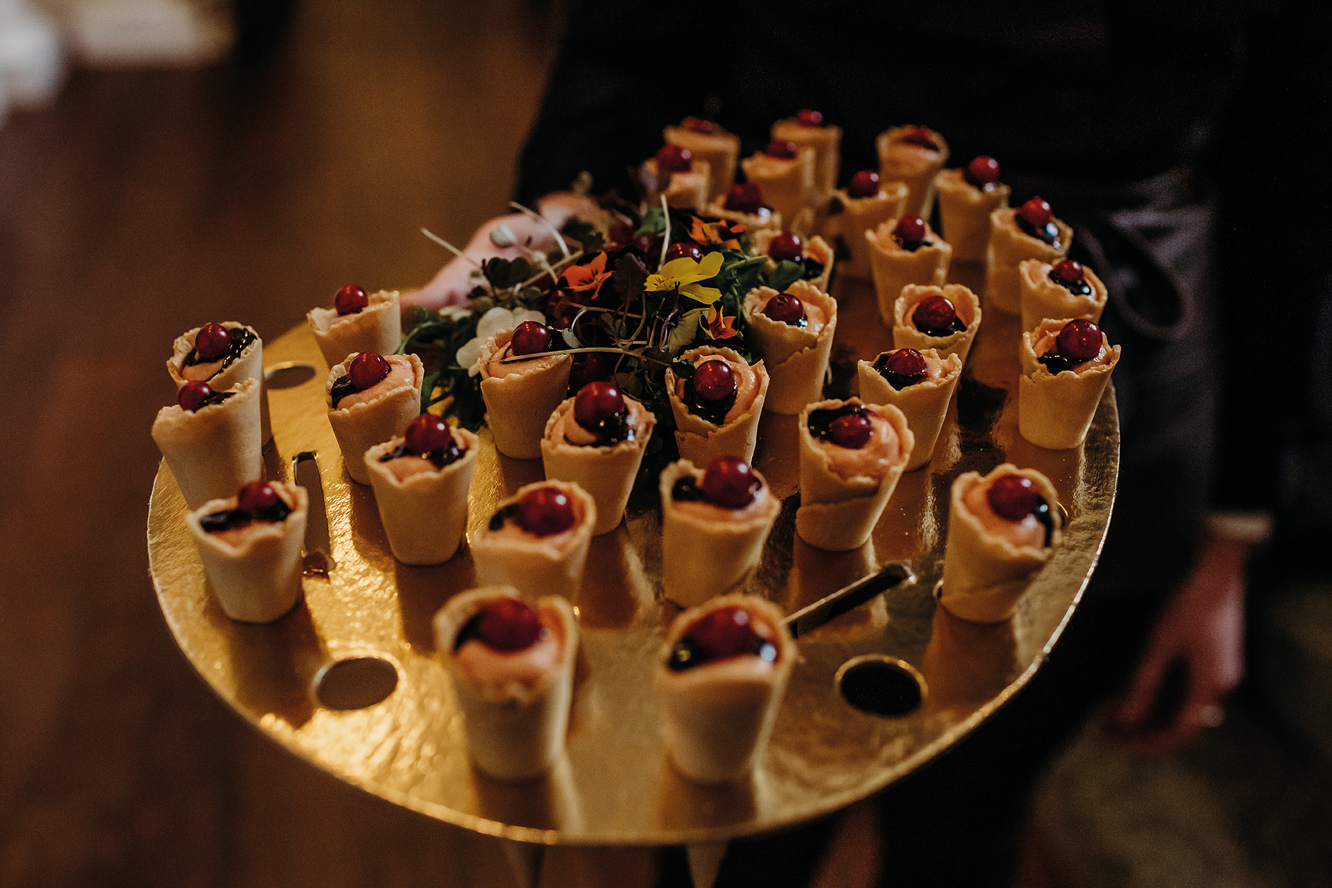 A tray of bite-sized appetizers in pastry cones, topped with cherry-like garnishes, presented on a gold platter.