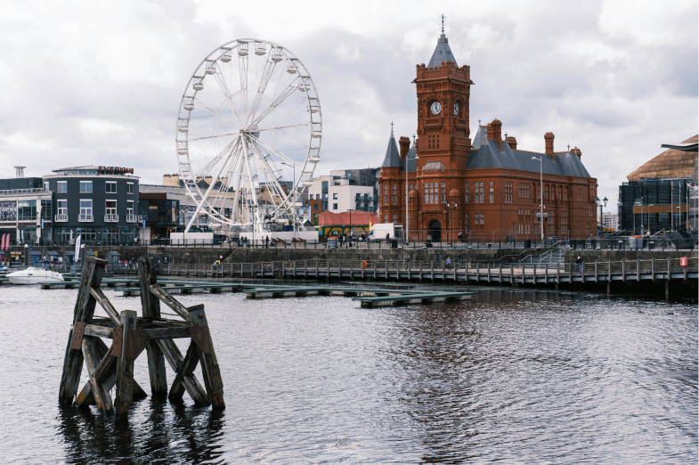 A waterfront scene with a Ferris wheel and a red brick building with a clock tower under a cloudy sky.