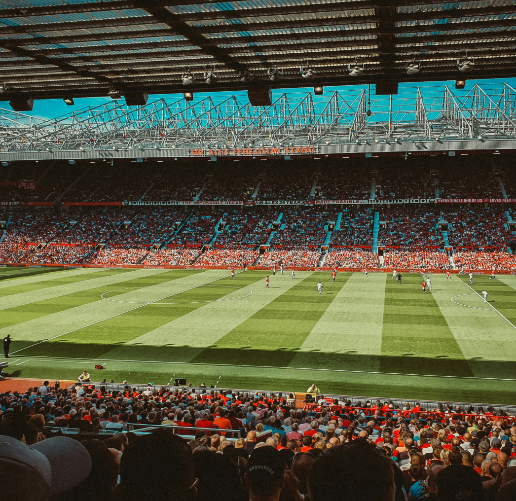 A wide-angle view of a soccer match in a large stadium filled with spectators. The field is green with players in action, and the sky is clear above.