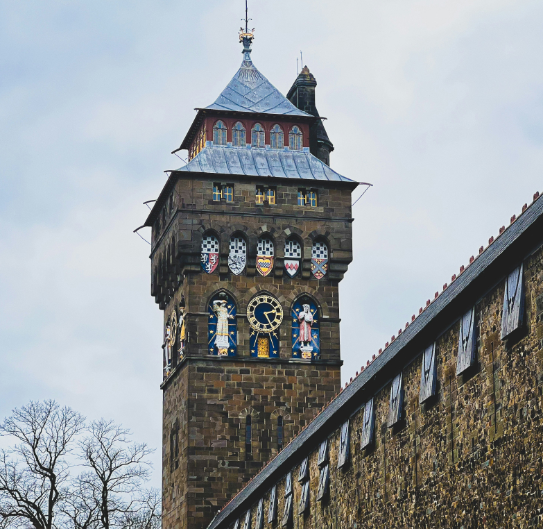 A stone clock tower with decorative shields and windows stands under a cloudy sky, alongside a tree-lined stone wall.