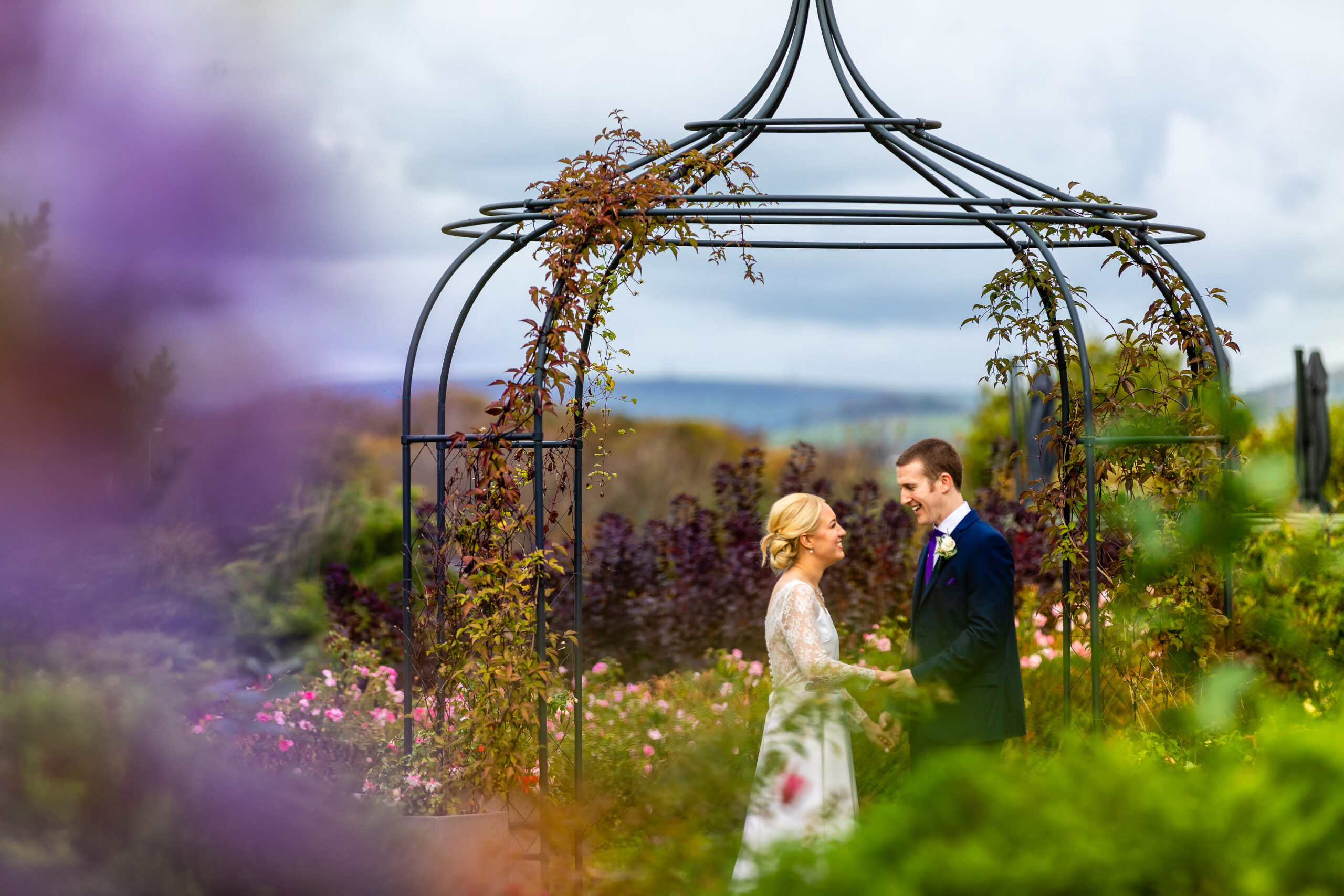 A couple standing and holding hands under a decorative arch in a garden setting.