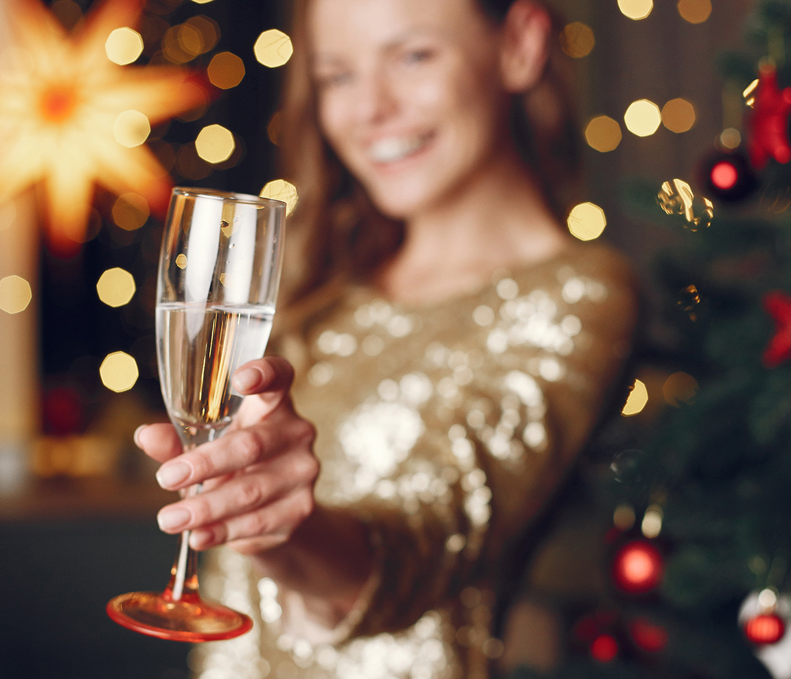 A woman in a sparkly dress holds a champagne flute, with a decorated Christmas tree and festive lights in the background.