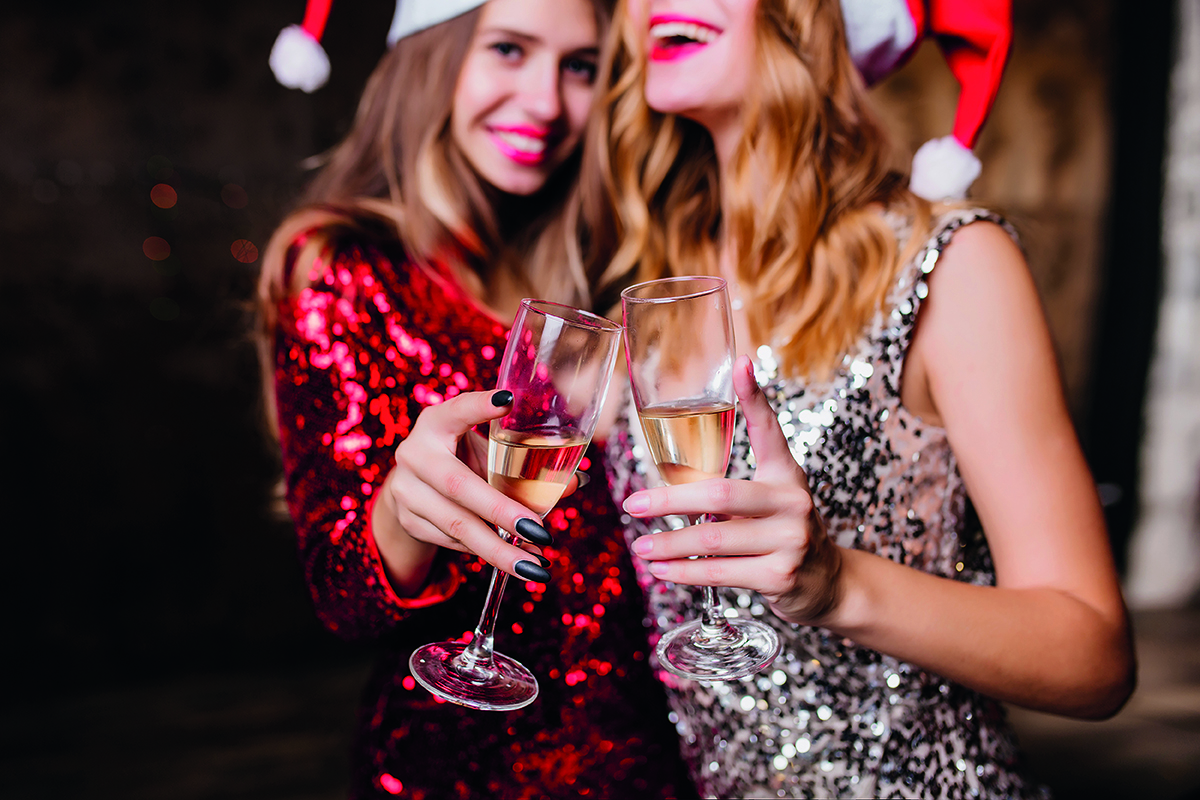 Two women in festive hats clinking champagne glasses, dressed in sequined outfits, smiling in a celebratory mood.